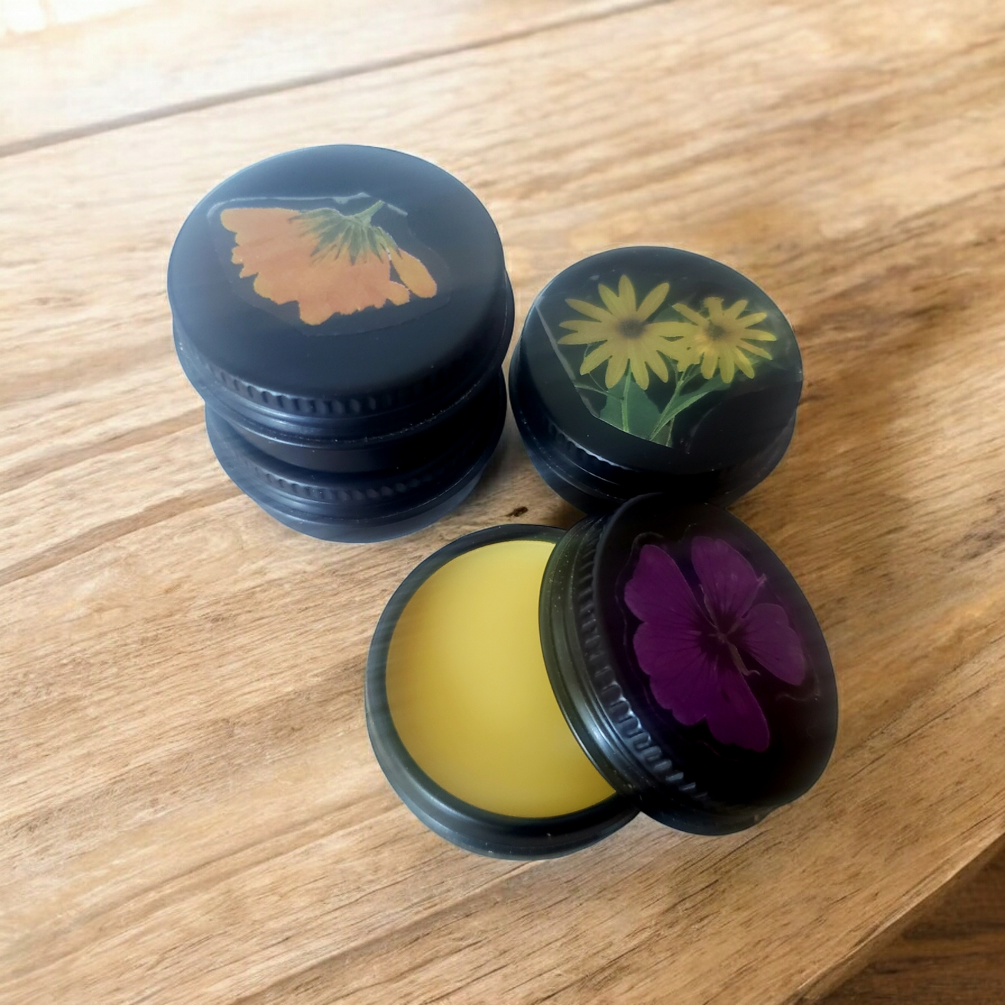 Three small, round containers with black lids, each featuring a different floral design on the top. One container is open, revealing a yellow beeswax balm inside.